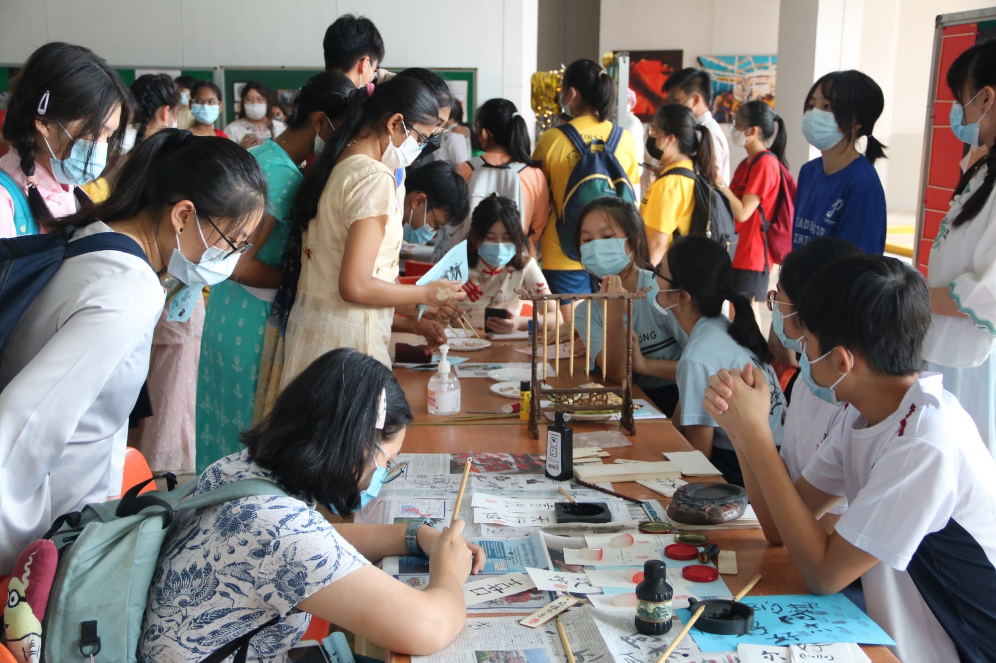 Students wearing face masks gather around a table doing Chinese calligraphy with ink and brushes at a school event.